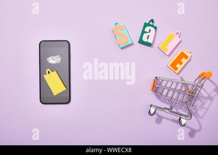 top view of smartphone near shopping bags with sale lettering falling into toy cart on violet, e-shopping concept Stock Photo