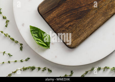 Top view of round marble surface with brown wooden cutting board and basil leaf near thyme twigs on white background Stock Photo