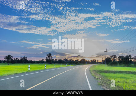 Silhouette the sunrise with the road, green paddy rice field, the beautiful sky and cloud in Thailand, by the beam, light and lens flare effect tone. Stock Photo