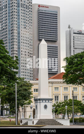 Singapore - March 20, 2019: White tall Dalhousie Obelisk on pedestal in front of skyscrapers of Financial District. Some green foliage at the fringes. Stock Photo