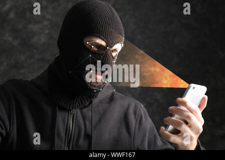 Male hacker in mask holding smartphone protected with facial recognition system Stock Photo