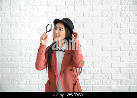 Female Asian detective with magnifying glass against white brick wall Stock Photo