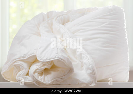 A duvet lies on a chest of drawers against a blurred window. Household. Stock Photo