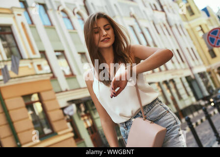 Portrait of young stylish woman walking on street, wearing cute trendy outfit. She is looking at her watch. Summer lifestyle concept. Horizontal shot Stock Photo