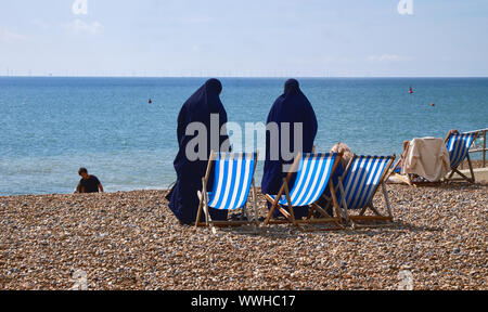 Two Muslim women standing on rocky Brighton beach about to sit down on deck chair next to their blonde hair friend as seen from top Stock Photo