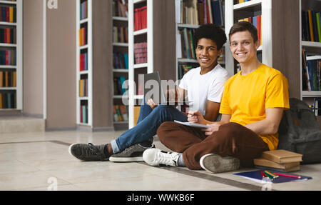 Two guys studying on floor in library, smiling at camera Stock Photo