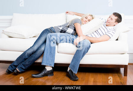Young couple recline on a white sofa with the young woman lying on the man with a pillow. Horizontal shot. Stock Photo