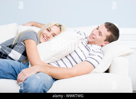 Young couple recline on a white sofa with the young woman lying on the man with a pillow. Horizontal shot. Stock Photo