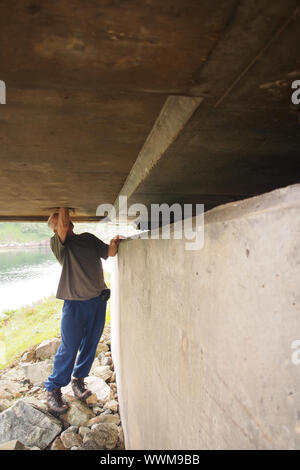 An older man under a concrete road bridge looking up with his hand and arm up to the elbow through a hole in the concrete Stock Photo