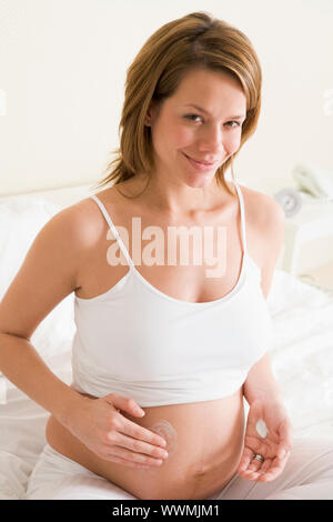 Pregnant woman in bedroom rubbing cream on belly smiling Stock Photo