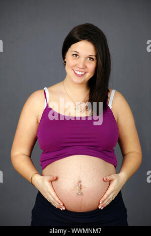 Studio shot portrait of a naturally beautiful, happy and healthy pregnant young woman holding her belly and smiling over grey background. Stock Photo