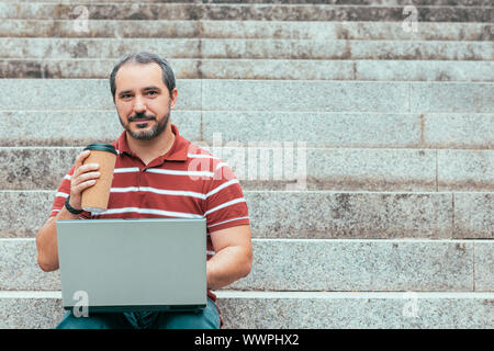man sitting on staircase outside and working on laptop. Taking coffee Stock Photo