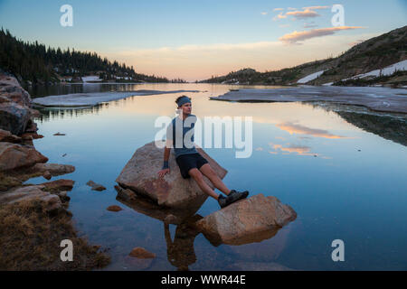 A man rests from a trail run at sunset sitting on boulders in Lake Isabelle, Indian Peaks Wilderness, Colorado. Stock Photo