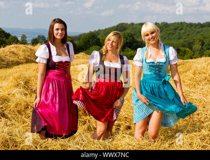 Three happy girls dancing in dirndls on field with straw Stock Photo