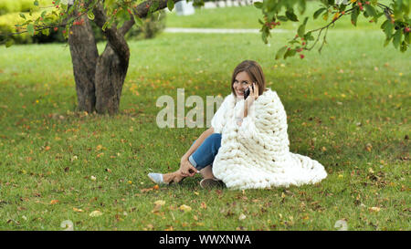 A young girl wrapped in a merino plaid talking on the phone sitting on the grass in a city park. Stock Photo