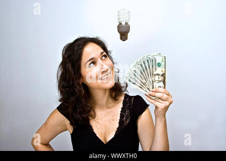 Woman waving money and looking up. Having an environmentally friendly idea with an energy saving fluorescent light bulb floating above her head, on a Stock Photo