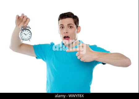 Shocked young guy holding alarm clock and pointing at it Stock Photo