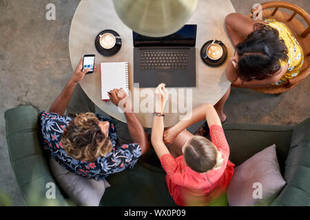 Candid overhead shot of three multi-ethnic millennial coworkers collaborating over coffee with laptop computer at bright cafe serving fair-trade Stock Photo