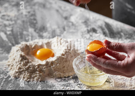 A women's hands holds a shell with yolk on the background of a kitchen table with flour. Preparation of dough for baking Stock Photo