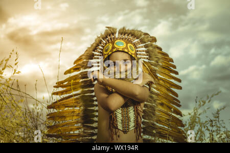 American Indian boy dressed in traditional feathered costume, wearing a tuft on his head, picture at sunset in a wheat field Stock Photo