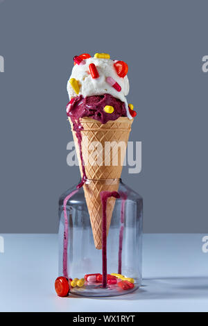 The waffle cone with a cold ice cream stands in glass jar with red and yellow capsules and a lollipop. Jar on a gray background. Stock Photo