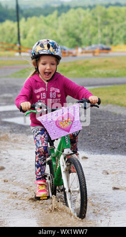 A smiling little girl (5 yrs old) riding her bike through a puddle Stock Photo