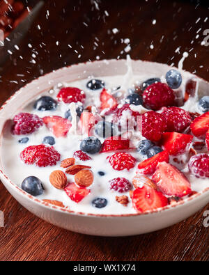 Splash milk from a bowl with a natural diet breakfast with granola, nuts and berries falling on a wooden table. Stock Photo
