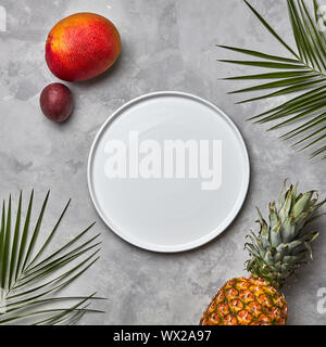 Composition from an empty white plate of green palm leaves, pineapple, mango and passion fruit on a gray marble table with a cop Stock Photo
