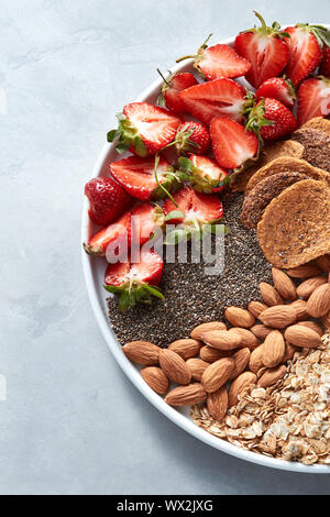 Homemade oat granola with almond nuts, slices of strawberries, seed chia, in a white plate on a gray background. Copy space. Top Stock Photo