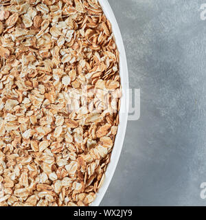 Uncooked oat flakes in a white bowl on a gray background. Close up, top view of natural organic product. Stock Photo