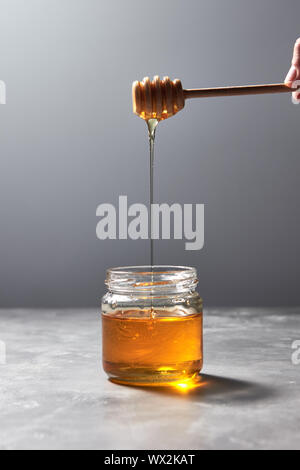 Woman hand hold a wooden stick with dripping sunflower honey in a glass jar on a gray background. Jewish New Year healthy holida Stock Photo