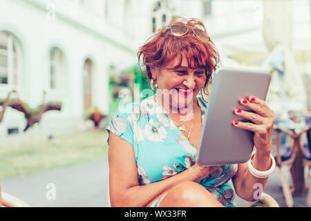 Smiling senior woman spending time in a outdoor cafe using digital tablet. Attractive senior woman  laughing while reading electronic book via digital Stock Photo