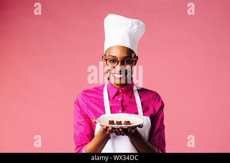 Portrait of a smiling young female chef holding a plate with three chocolate pralines, isolated on pink studio background Stock Photo