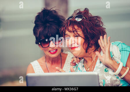 Two happy senior women - friends waving hand and talking while having a video call with their friends on a laptop in a cafe. Stock Photo