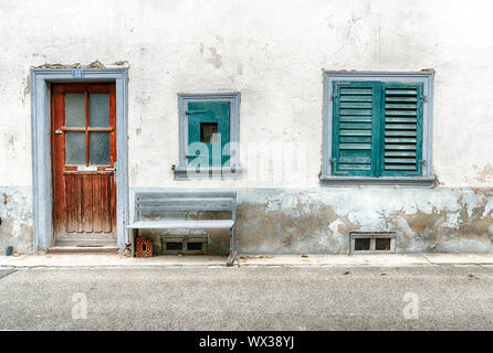 worn and faded house front detail with two windows and old wooden door and rickety bench on the road Stock Photo