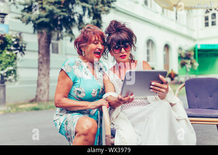 Two senior women sitting in outdoor cafe laughing and watching social media and funny viral videos on a tablet. Stock Photo