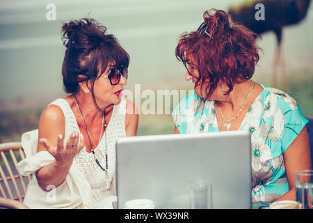 Two happy senior women talking and using laptop on a laptop in outdoor cafe. Stock Photo