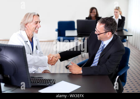 Mature doctor smiling and shaking hands with a patient at his desk in his office - colleagues in the background Stock Photo