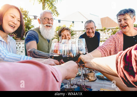 Happy senior friends toasting with red wine glasses at dinner on patio - Mature people having fun dining together outside