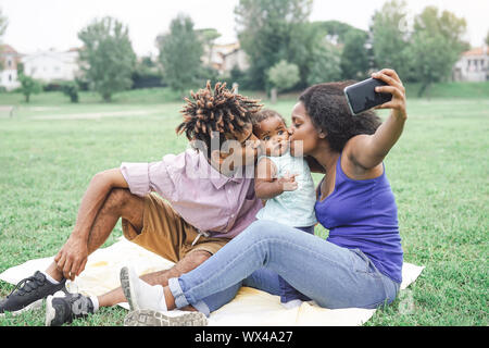 Happy african family taking a selfie with mobile smart phone camera in a public park outdoor - Mother and father having fun with their daughter Stock Photo