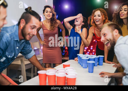 Happy friends playing beer pong in a cocktail bar - Young millennial people having fun doing party alcohol games at night pub Stock Photo