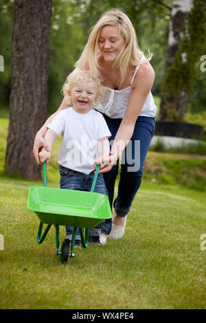 A mother playing with her son pushing a wheelbarrow outdoors Stock Photo