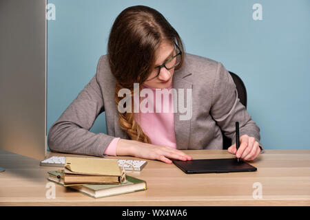 A girl in a pink sweater, gray jacket and glasses sits at a table, bowing her head, draws on an electronic tablet. Stock Photo