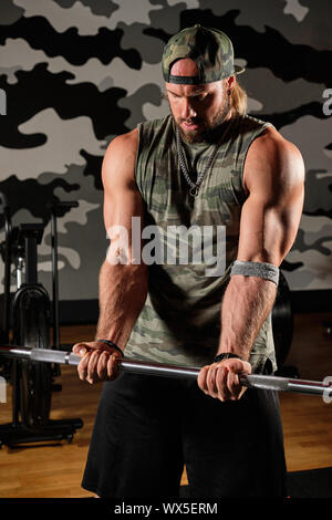 Muscular man in a military t-shirt with a print and a baseball cap holds the barbell in his hands, his head down Stock Photo