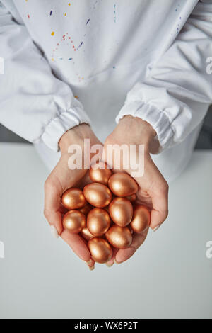 Golden painted small eggs hold the hands of a woman on a gray background. Concept of profit and wealth. Top view Stock Photo