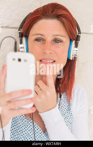 Pretty smiling redhead woman sitting against an old wall the finger under the mouth listening to music on her phone with a headset Stock Photo