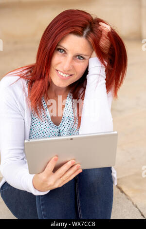 Portrait of a beautiful red-haired woman with a serene face and hand in hair using a tablet Stock Photo