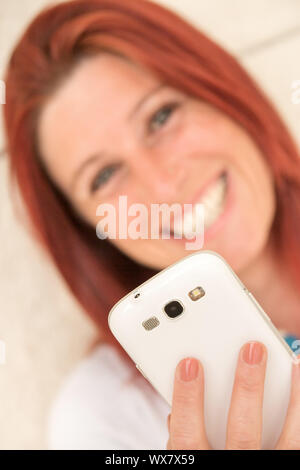 Pretty smiling redhead woman sitting against an old wall isending messages or make a selfie with her mobile phone. Focus on mobile phone Stock Photo