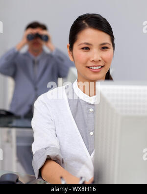 Self-assured asian businesswoman at a computer with her colleague using binoculars in the background Stock Photo