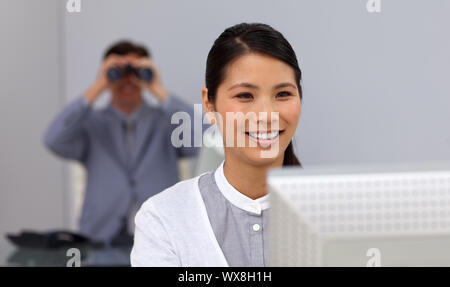 Smiling businesswoman at a computer with her colleague using binoculars in the background Stock Photo